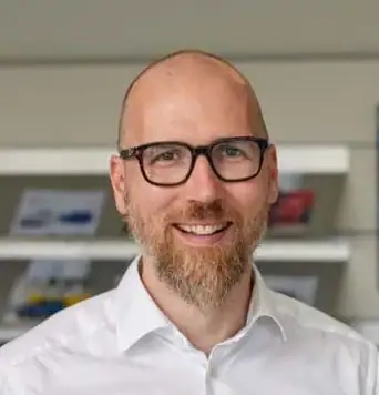 Ein glatzköpfiger Mann mit Brille und Bart lächelt in einem weißen Hemd bei der Ring Garage AG, hinter ihm stehen Regale. Ein glatzköpfiger Mann mit Brille und Bart lächelt in einem weißen Hemd bei der Ring Garage AG, hinter ihm stehen Regale.