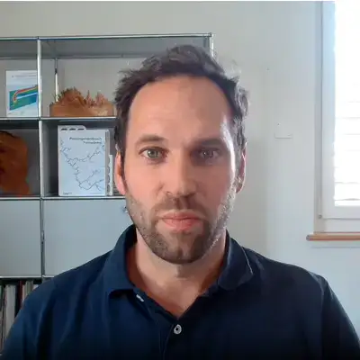 Ein Mann mit kurzen Haaren und Bart sitzt in einem Raum vor Regalen und einem Fenster. Ein Mann mit kurzen Haaren und Bart sitzt in einem Raum vor Regalen und einem Fenster.